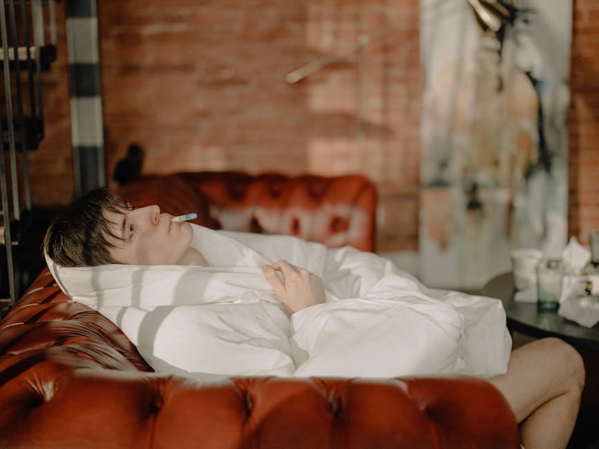 A young man wrapped in a blanket with a thermometer, relaxing on a couch in a cozy home setting.